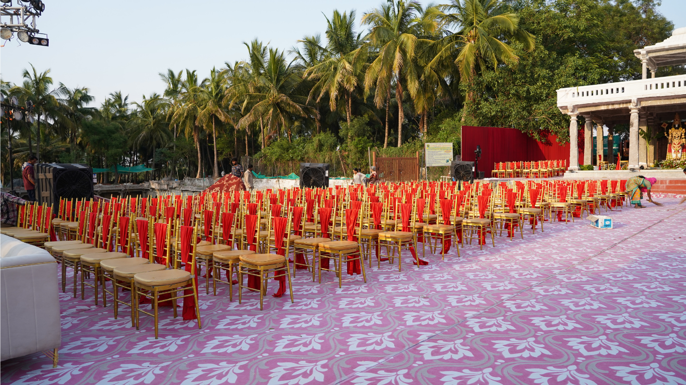 Guest seating with red-draped gold chairs under palm trees beside the temple mandap